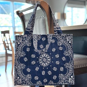 May include: A navy blue quilted tote bag featuring a white paisley bandana print pattern. The bag has two matching fabric handles and vertical stitching lines across the front, showcasing a classic repeating floral and geometric design.