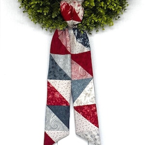 May include: A long, narrow fabric scarf featuring a patchwork design of triangular segments. The pattern consists of various floral and paisley prints in shades of deep red, slate blue, and cream, arranged in a repeating geometric layout.