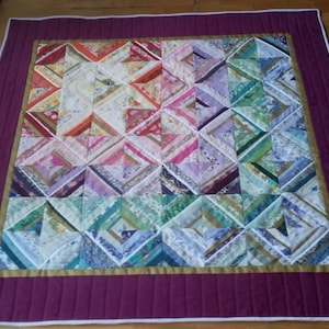 May include: A colorful quilt with a geometric pattern made from strips of fabric in shades of pink, purple, green, blue, and gold. The quilt is bordered with a wide burgundy band and a thin white band.