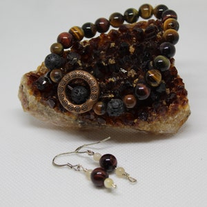 Puede incluir: Un brazalete hecho con ojo de tigre, roca volcánica y un dije de cobre. El brazalete se muestra en un cúmulo de cristales. También se muestran pendientes a juego con ojo de tigre, blanco y cuentas de cobre.
