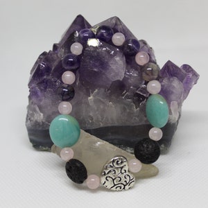 Op de afbeelding: Een armband gemaakt van amethist, rozenkwarts, turquoise, lavasteen en een zilveren hartbedel. De armband wordt getoond op een groot amethistkristal.