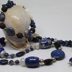 Puede incluir: Un conjunto de joyas de lapislázuli que incluye un collar, una pulsera y pendientes. El collar y la pulsera están hechos con cuentas de lapislázuli, cuentas doradas y cuentas de roca volcánica negra. Los pendientes están hechos con cuentas de lapislázuli y ganchos de plata.