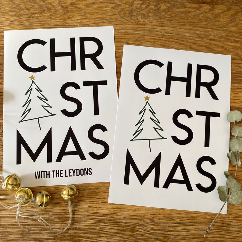K&ouml;nnte beinhalten: Zwei wei&szlig;e rechteckige Drucke mit dem schwarzen Schriftzug CHRISTMAS, bei dem ein gezeichneter Weihnachtsbaum den Buchstaben I ersetzt. Ein Druck enth&auml;lt den schwarzen Text WITH THE LEYDONS. Goldene Gl&ouml;ckchen und ein Eukalyptuszweig liegen dabei.
