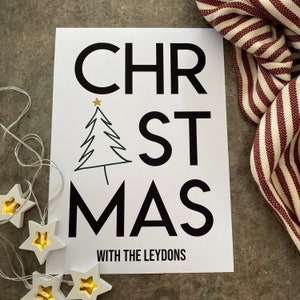 K&ouml;nnte beinhalten: Ein wei&szlig;er rechteckiger Druck mit dem schwarzen Schriftzug CHRISTMAS, bei dem ein gr&uuml;ner, liniengezeichneter Weihnachtsbaum den Buchstaben I ersetzt und von einem goldenen Stern gekr&ouml;nt wird. Darunter steht WITH THE LEYDONS in Schwarz, neben sternf&ouml;rmigen Lichtern.