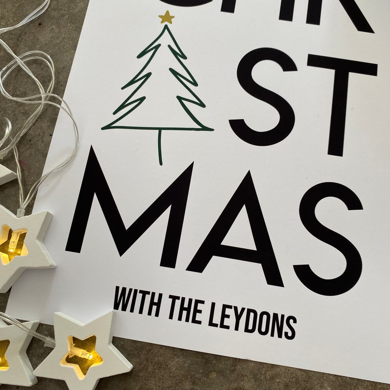 K&ouml;nnte beinhalten: Ein wei&szlig;es rechteckiges Schild mit fettem schwarzem Text CHRISTMAS WITH THE LEYDONS und einem gr&uuml;nen Strichzeichnungs-Weihnachtsbaum mit goldenem Stern. Eine Lichterkette mit wei&szlig;en Sternen und warmgelbem Licht liegt links daneben.