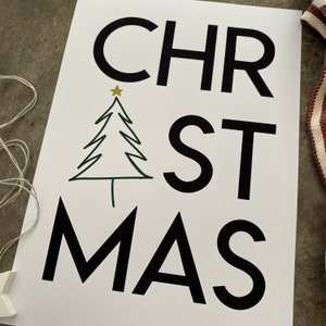 K&ouml;nnte beinhalten: Ein wei&szlig;es rechteckiges Schild mit dem schwarzen Schriftzug CHRISTMAS, bei dem der Buchstabe I durch eine gr&uuml;ne Strichzeichnung eines Tannenbaums mit einem kleinen goldenen Stern an der Spitze ersetzt ist. Das Schild liegt neben wei&szlig;en Lichterketten.