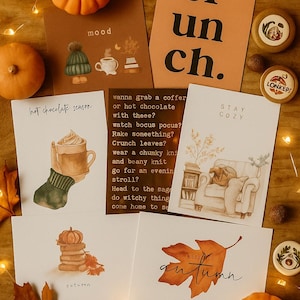 May include: A seasonal arrangement with illustrated greeting cards, pumpkins, and decorative items. The cards feature text and illustrations, including phrases like "hot chocolate season" and "autumn". The image has a warm colour scheme with orange, brown, and cream tones.