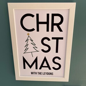 K&ouml;nnte beinhalten: Ein wei&szlig; gerahmter Wanddruck mit dem schwarzen Schriftzug "CHRISTMAS", bei dem ein stilisierter schwarzer Strichzeichnungs-Weihnachtsbaum den Buchstaben "I" ersetzt und von einem kleinen goldenen Stern gekr&ouml;nt wird. Darunter steht "WITH THE LEYDONS".
