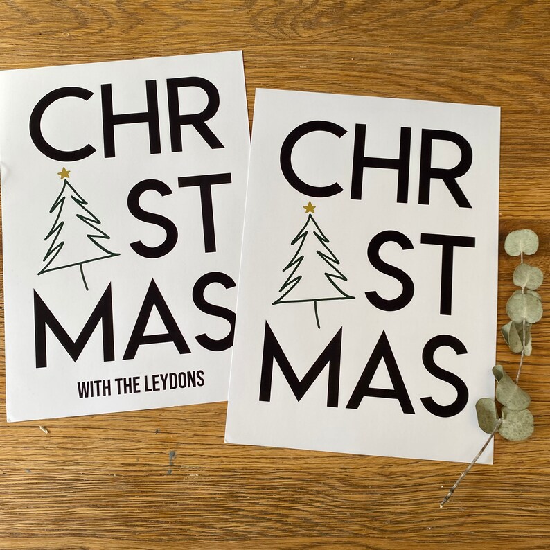 K&ouml;nnte beinhalten: Zwei wei&szlig;e rechteckige Drucke mit dem schwarzen Text CHRISTMAS in einer fetten serifenlosen Schrift. Ein einfacher schwarzer Strichzeichnungs-Weihnachtsbaum mit einem kleinen goldenen Stern ersetzt den Buchstaben I. Ein Druck enth&auml;lt den Text WITH THE LEYDONS.