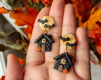 Könnte beinhalten: Ein Paar Halloween-Ohrringe. Jeder Ohrring zeigt ein schwarzes Haus mit einem Kürbis und einem Mond mit Fledermäusen. Die Ohrringe werden in einer Hand gehalten, mit Herbstlaub im Hintergrund.
