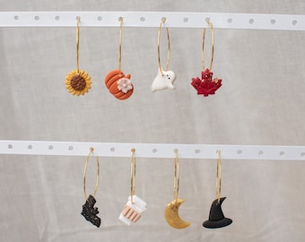 Könnte beinhalten: Ein Set mit acht goldenen Creolen mit verschiedenen Herbst- und Halloween-Anhängern. Die Anhänger umfassen eine Sonnenblume, einen Kürbis, ein Gespenst, ein Ahornblatt, eine Fledermaus, eine Kaffeetasse, einen Halbmond und einen Hexenhut.