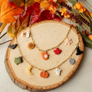 Puede incluir: Dos collares dorados con colgantes temáticos de otoño y Halloween. Los colgantes incluyen una calabaza, una hoja de arce, un fantasma, un sombrero de bruja y un girasol. Los collares descansan sobre una rodaja de madera con follaje otoñal.