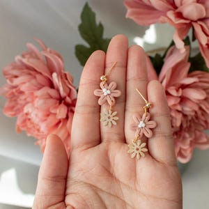 Pode incluir: Um par de brincos de argola dourados com dois pingentes de flores cor-de-rosa e um pingente de flor dourada menor em cada brinco.