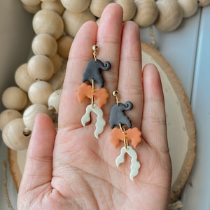 May include: A pair of dangle earrings featuring three clay shapes: a black elephant, an orange swirl, and a white swirl. The earrings have gold hooks.