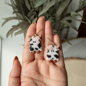 May include: A pair of cow-shaped earrings with black and white spots and a pink flower accent. The earrings are made of polymer clay and have gold hooks.