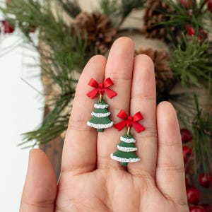 May include: A pair of Christmas tree earrings. Each earring features a green tree with white trim and a red bow at the top. The earrings are held in a hand, with a blurred background of greenery and pine cones.