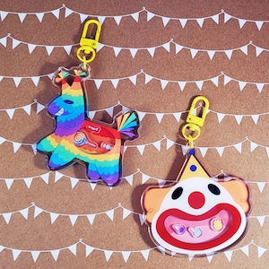 May include: Two acrylic keychains with yellow clasps. One is a colorful piñata with candy inside. The other is a clown face with a red nose and a yellow party hat. The background is brown with white pennant banners.