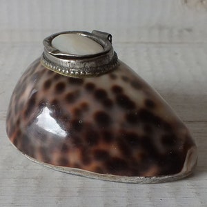 Op de afbeelding: Een klein, bruin en zwart schelpje met een zilveren deksel en een witte parelmoer inleg.