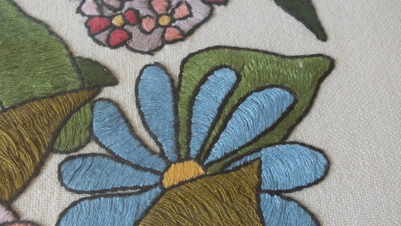 May include: Close-up of an embroidered blue flower with a yellow centre, surrounded by green leaves. The flower is stitched with a detailed, textured design.