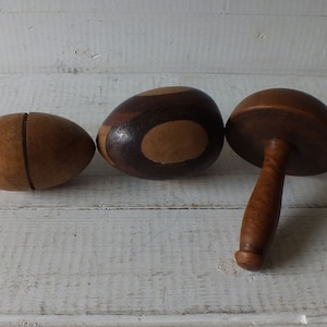 Op de afbeelding: Drie houten eivormige ornamenten, een met een bruin en tan swirl-patroon, een met een bruin en tan gesplitst ontwerp en een met een bruine paddenstoelvormige bovenkant en een bruine handgreep.