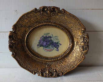May include: An oval frame with ornate gold detailing surrounds a cross-stitch embroidery of a basket of purple flowers.