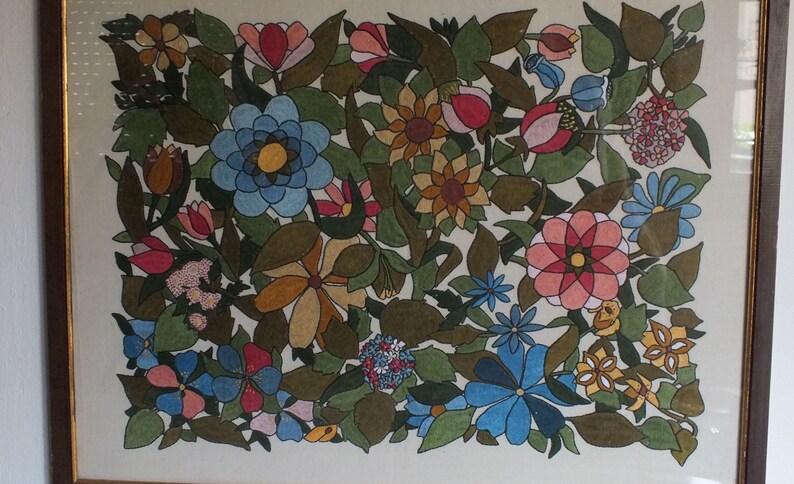 May include: A framed artwork featuring a colourful floral design with various flowers, leaves, and stems. The flowers are in shades of pink, red, blue, yellow, and brown. The background is a light cream colour.