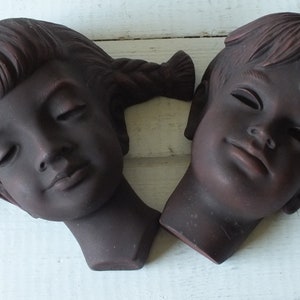 May include: Two ceramic wall sculptures of children's faces. The sculptures are brown and have a textured finish. The girl's sculpture has her hair in pigtails and her eyes are closed. The boy's sculpture has his hair parted on the side and his eyes are closed.