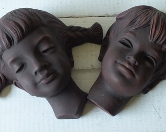 May include: Two ceramic wall sculptures of children's faces. The sculptures are brown and have a textured finish. The girl's sculpture has her hair in pigtails and her eyes are closed. The boy's sculpture has his hair parted on the side and his eyes are closed.
