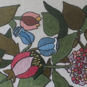 May include: Embroidered floral design featuring pink, red, blue, and green flowers on a white background. The flowers are surrounded by green leaves.