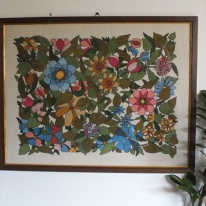 May include: A framed artwork featuring a colourful floral design. The artwork is painted in a variety of colours, including red, pink, blue, yellow, and green. The flowers are arranged in a symmetrical pattern, and the background is a light cream colour.