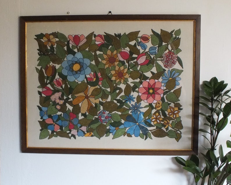 May include: A framed artwork featuring a colourful floral design. The artwork is painted in a variety of colours, including red, pink, blue, yellow, and green. The flowers are arranged in a symmetrical pattern, and the background is a light cream colour.