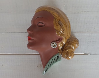 May include: A ceramic wall hanging depicting a woman's profile. The face is a dark brown with red lips and closed eyes. The hair is light brown, styled in a bun. The earring is white with a gold centre. The neck has a green collar.