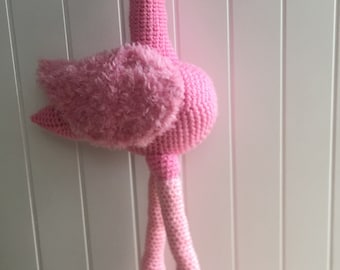 May include: A pink crocheted flamingo toy with a fluffy pink wing and black eyes. The flamingo is standing on a white surface.
