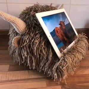 May include: A novelty tablet stand in the form of a Highland cow, made from brown yarn. The stand has a shaggy texture, small ears, and a single horn. A white tablet displays an image of a Highland cow. The stand is designed to hold the tablet upright.