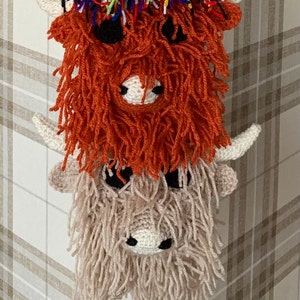 May include: Two crocheted Highland cow ornaments hanging on a plaid background. The top cow is orange with black eyes and a white nose. The bottom cow is beige with black eyes and a white nose.
