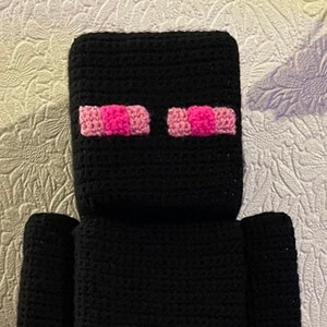 May include: Black crocheted plush toy with pink eyes. The toy is shaped like a character from a popular video game.