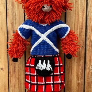 May include: A crocheted stuffed toy Highland cow wearing a blue and white Scottish kilt and a blue and white Scottish flag shirt. The toy is standing on a blue surface.