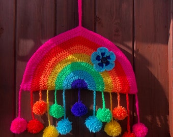 May include: A handmade crochet rainbow wall hanging. The rainbow features vibrant colors including pink, orange, yellow, green, and blue. It is adorned with a blue flower and colorful pom-poms hanging below. The wall hanging is suspended by a pink loop.