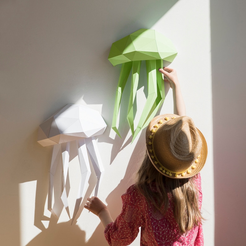 May include: A child wearing a straw hat with yellow pom-poms decorates a wall with two paper jellyfish, one green and one white.