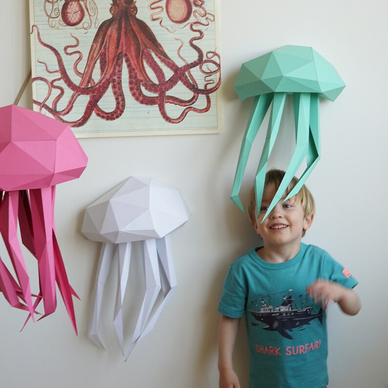 May include: Three papercraft jellyfish, one pink, one white, and one teal, are hanging on a white wall. A young child is standing in front of the teal jellyfish, smiling and looking at the camera. The teal jellyfish is hanging above the child's head.