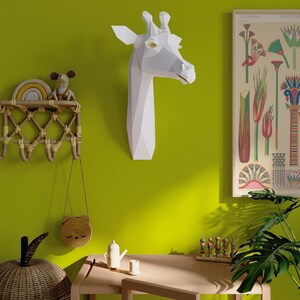 May include: A white paper giraffe head sculpture mounted on a green wall. A small wooden table with a wooden toy and a teapot sits in front of a potted plant.