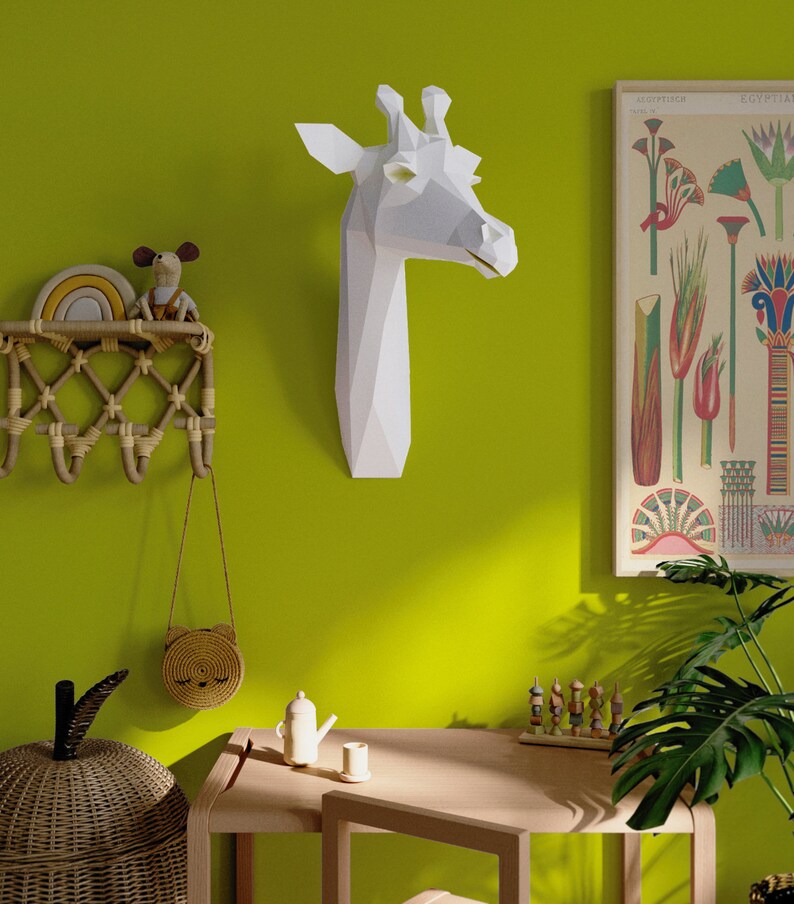 May include: A white paper giraffe head sculpture mounted on a green wall. A small wooden table with a wooden toy and a teapot sits in front of a potted plant.