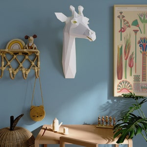 May include: A white paper giraffe head mounted on a light blue wall. A wooden table with a small teapot and a cup sits in front of a potted plant. A wicker basket and a small woven bag hang on a wall rack.