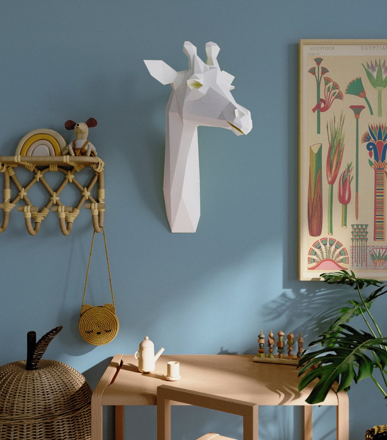 May include: A white paper giraffe head mounted on a light blue wall. A wooden table with a small teapot and a cup sits in front of a potted plant. A wicker basket and a small woven bag hang on a wall rack.