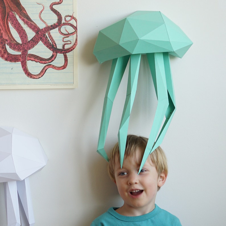 May include: A child wearing a teal blue papercraft jellyfish hat. The jellyfish has a geometric design and is made of paper.