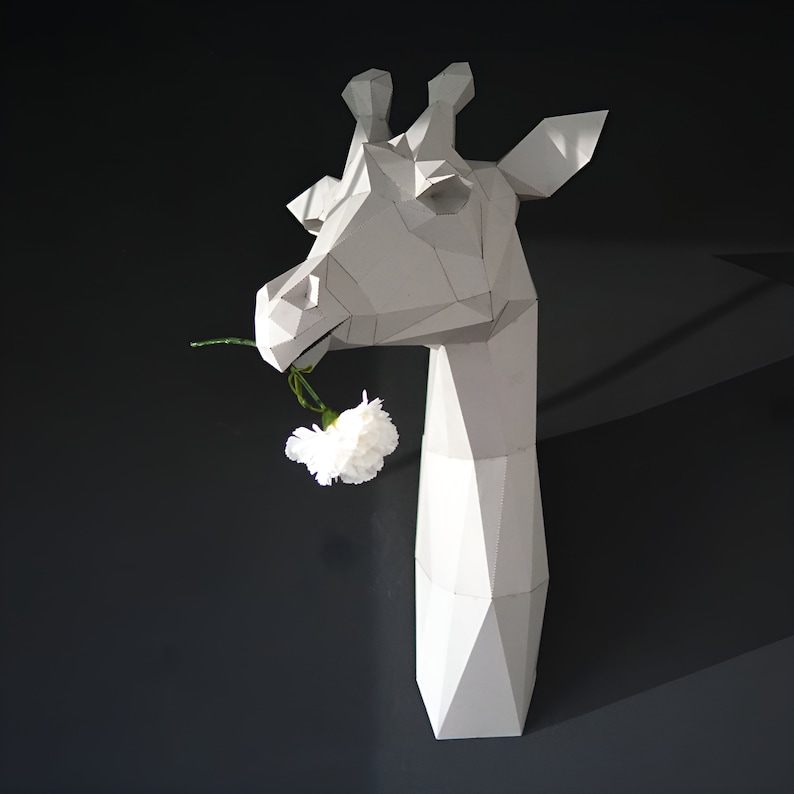 May include: A white papercraft giraffe head sculpture with a white carnation flower in its mouth.