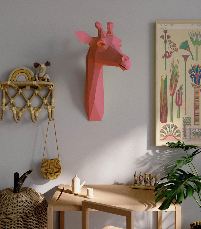 May include: A pink origami giraffe head mounted on a light gray wall, a wooden table with a small teapot and a cup, a wicker basket, a small wooden toy, and a potted plant with green leaves.