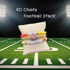 Könnte beinhalten: Drei gestapelte Armbänder mit Fußball-Motiv. Das obere Armband hat goldene Perlen und rote und gelbe Akzente. Das mittlere Armband zeigt einen Football-Anhänger. Das untere Armband schreibt "CHIEFS". Der Text "KC Chiefs Football Stack" steht oben.