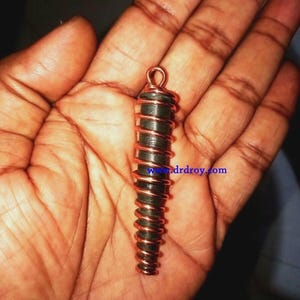 Puede incluir: Un colgante cilíndrico verde oscuro envuelto en alambre de cobre, sostenido en una mano abierta. El colgante tiene un lazo en la parte superior para colgar. La dirección del sitio web "www.drdroy.com" es visible en el colgante.