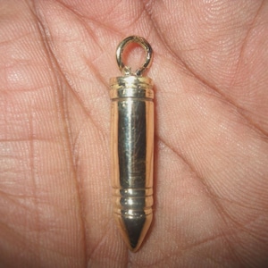 May include: A gold-colored bullet-shaped pendant with a loop at the top. The pendant has a cylindrical body with three horizontal grooves and a pointed tip. The pendant is held in a hand.